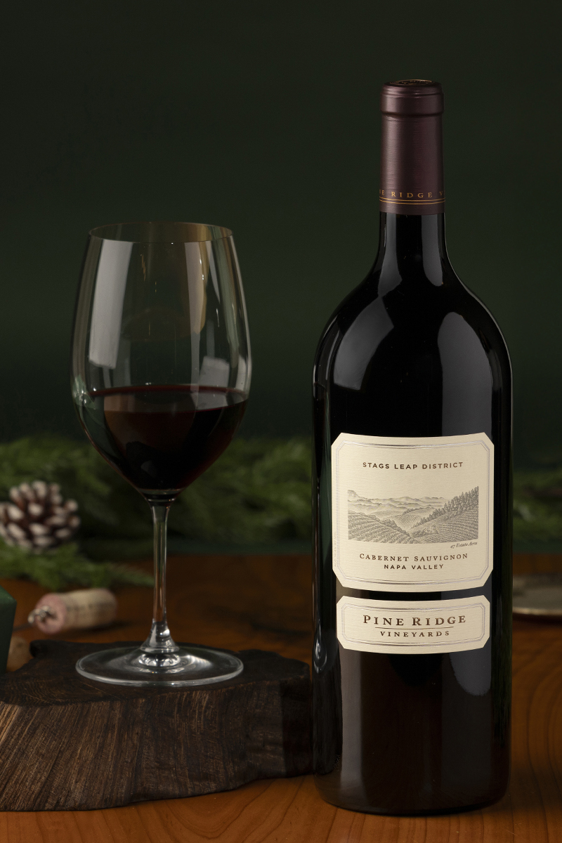 A Magnum of Pine Ridge Vineyards Stags Leap District Cabernet Sauvignon sits on a wooden surface beside a filled wine glass. Pine cones and lush greenery adorn the background, with a cork resting nearby.