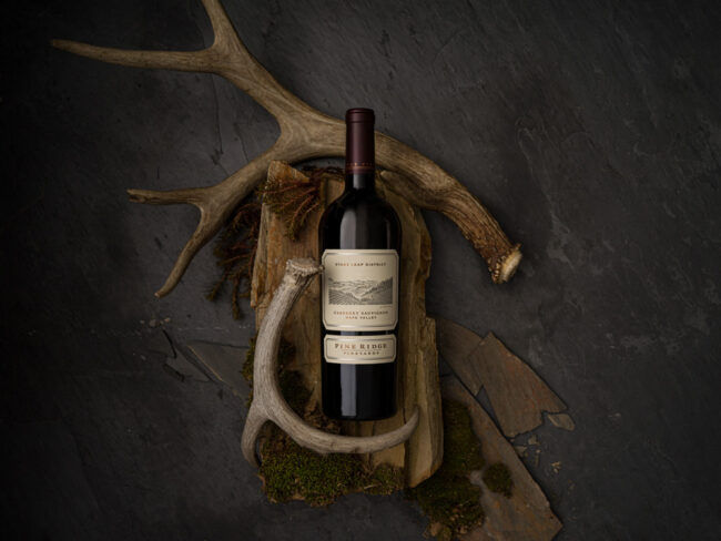 A bottle of Pine Ridge wine is placed horizontally on a dark slate surface. It is surrounded by two antlers, pieces of natural wood, and green moss, creating a rustic and earthy setting.