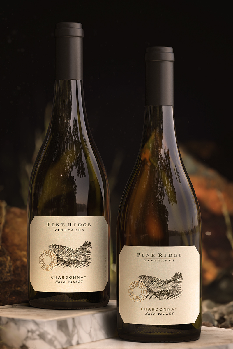 Two bottles of Pine Ridge Vineyards Chardonnay from Napa Valley are elegantly displayed. Each Chardonnay bottle boasts a beige label adorned with a vineyard illustration and is topped with a black screw cap, all set against a softly blurred natural background.