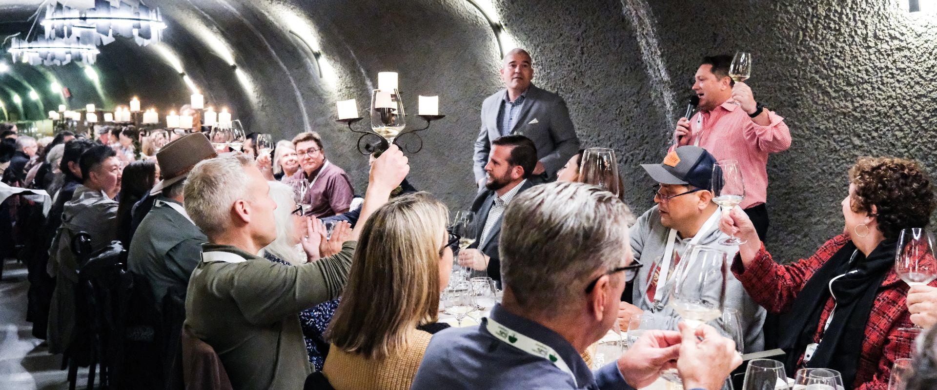 A group of people sits along a long table in an arched room, raising glasses in a toast. Two men are standing, one speaking into a microphone, while others listen attentively. The room is dimly lit by chandeliers, creating an intimate atmosphere.