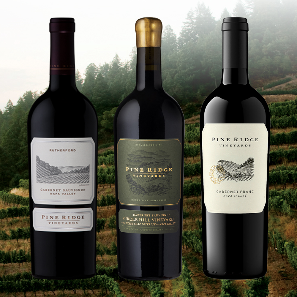 Three bottles of Pine Ridge Vineyards wine stand in a row against a blurred vineyard background, showcasing Rutherford and Circle Hill Cabernet Sauvignon and our Cabernet Franc.