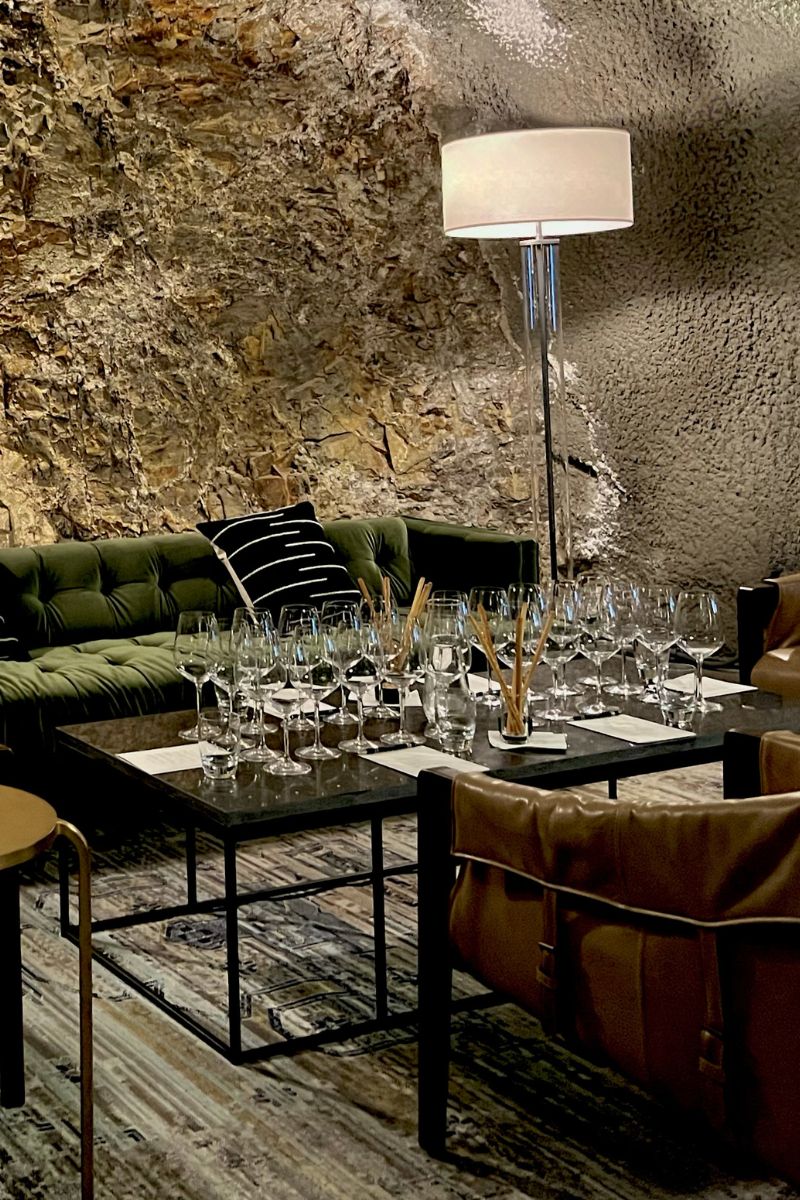A cozy room with a stone wall features a green sofa and several armchairs around a low table. The table is set with numerous wine glasses and breadsticks. A floor lamp with a white shade provides soft lighting.