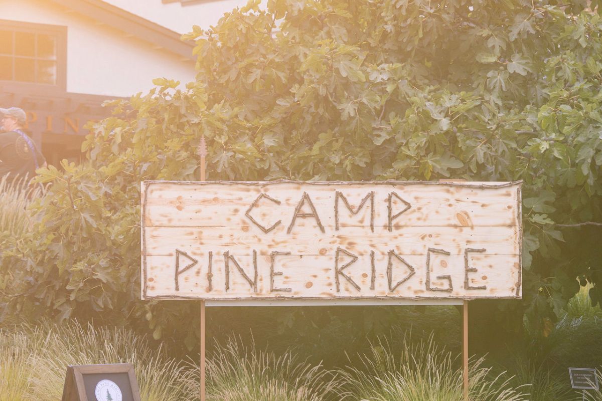 A rectangular wooden sign with the words Camp Pine Ridge carved into it stands outdoors in front of leafy green bushes and plants. The sign is held up by two posts.