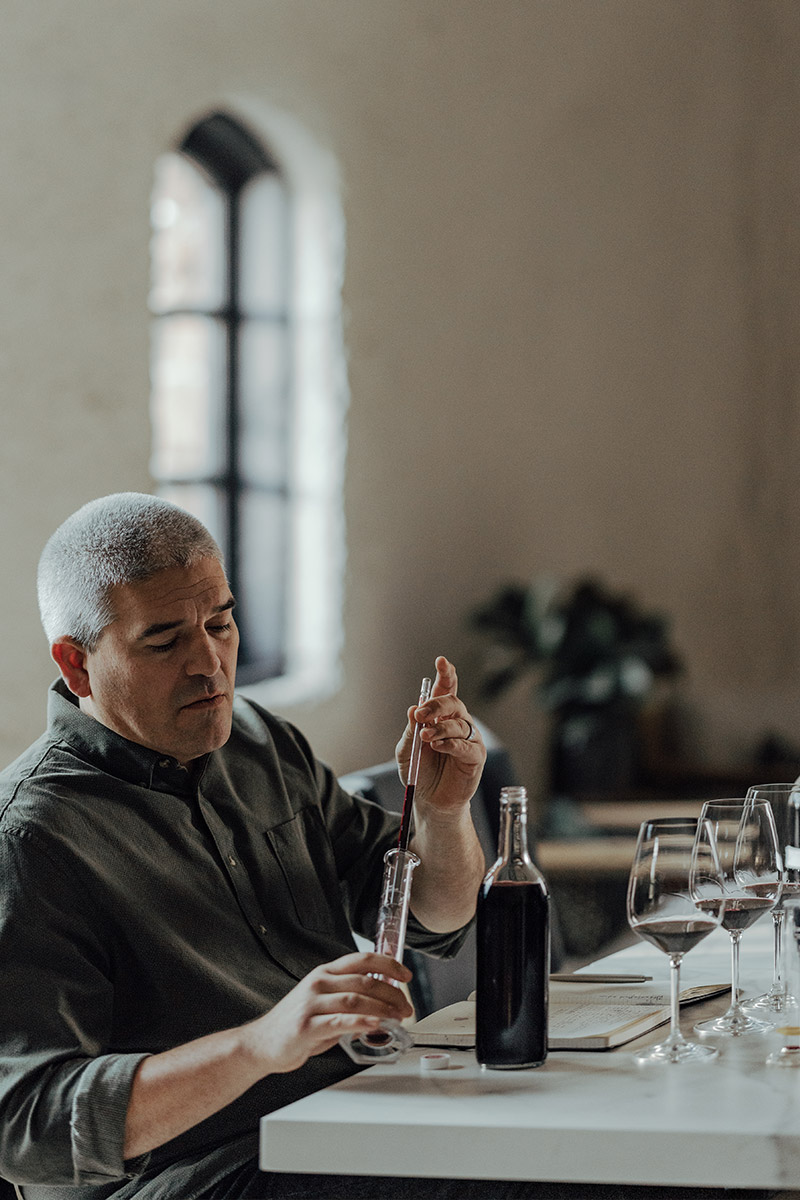A man with short gray hair uses a pipette to transfer red wine from a bottle into a glass. He is seated at a table with three empty wine glasses and a notebook. A window and a blurred plant are visible in the background.