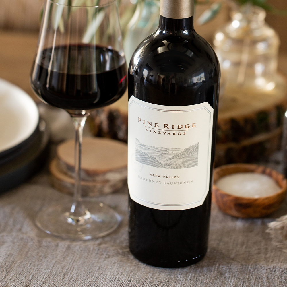 A bottle of Pine Ridge Vineyards Napa Valley Cabernet Sauvignon stands on a table next to a filled wine glass, a small bowl of salt, and other dining items, with a neutral tablecloth underneath.