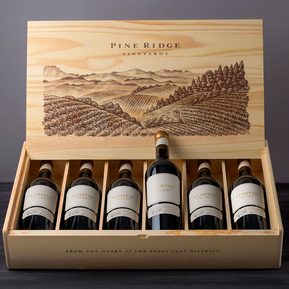 A wooden box labeled Pine Ridge Vineyards holds six bottles of red wine, with one bottle upright and five lying down. The box lid displays an engraved vineyard landscape scene.