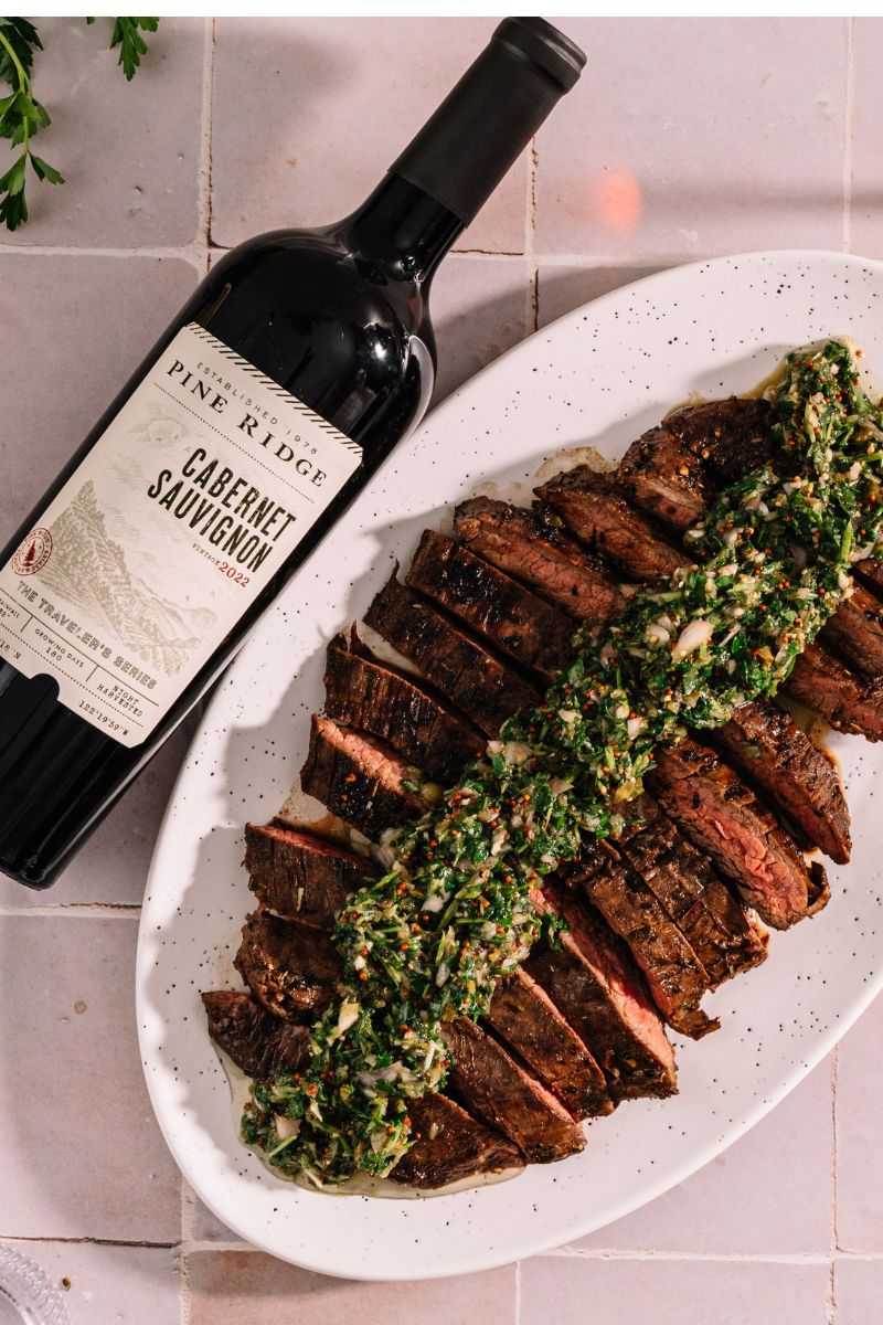 A sliced cooked steak on a white oval plate is topped with a green herb sauce. Next to the plate is a bottle of Pine Ridge Cabernet Sauvignon, placed on a light-colored tiled surface.
