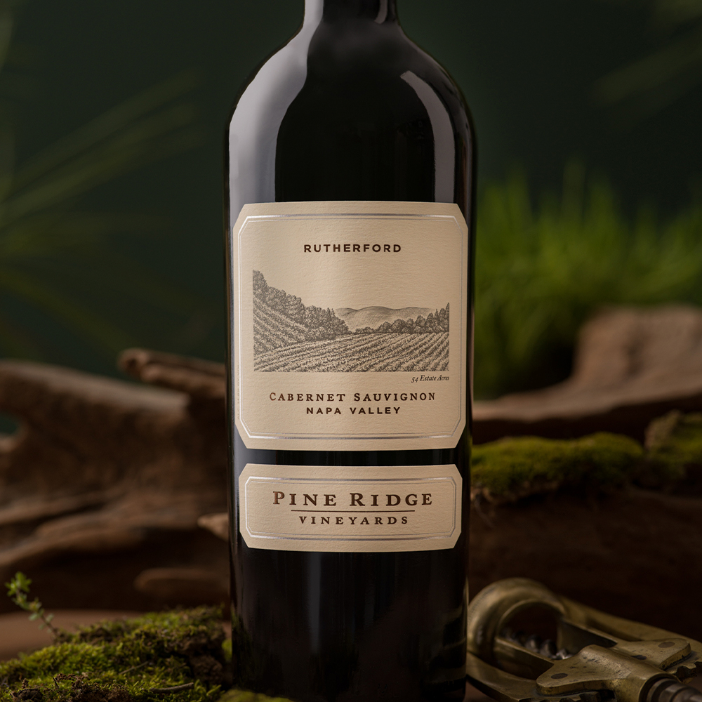 A bottle of Pine Ridge Vineyards Cabernet Sauvignon from the 2022 Rutherford Cabernet 3-Bottle Collection is displayed upright on a mossy surface with a blurred green background and a corkscrew partially visible beside it.