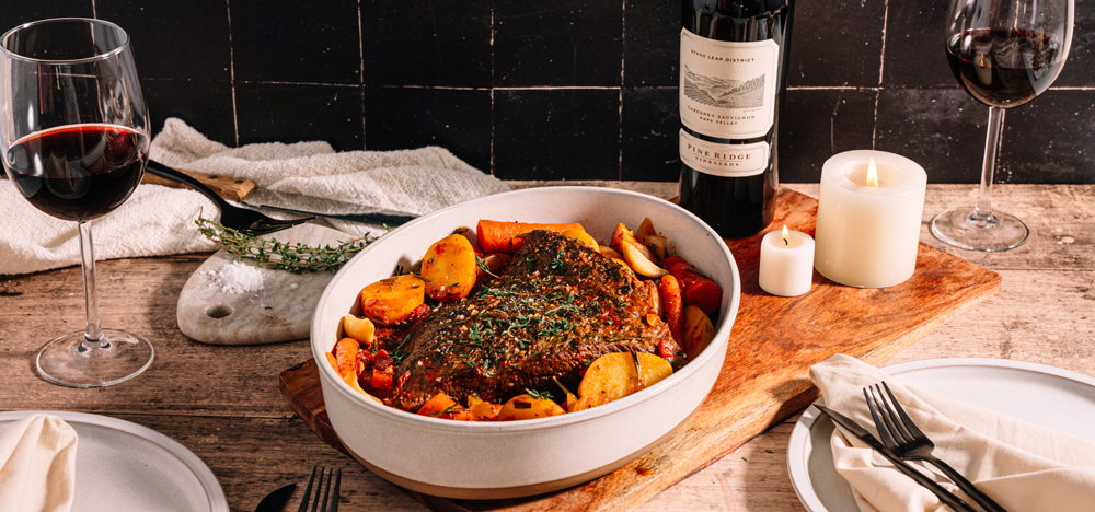 A white oval dish with cooked meat, potatoes, and vegetables sits on a wooden table beside two glasses of red wine, a bottle of wine, white candles, and place settings with napkins and utensils.