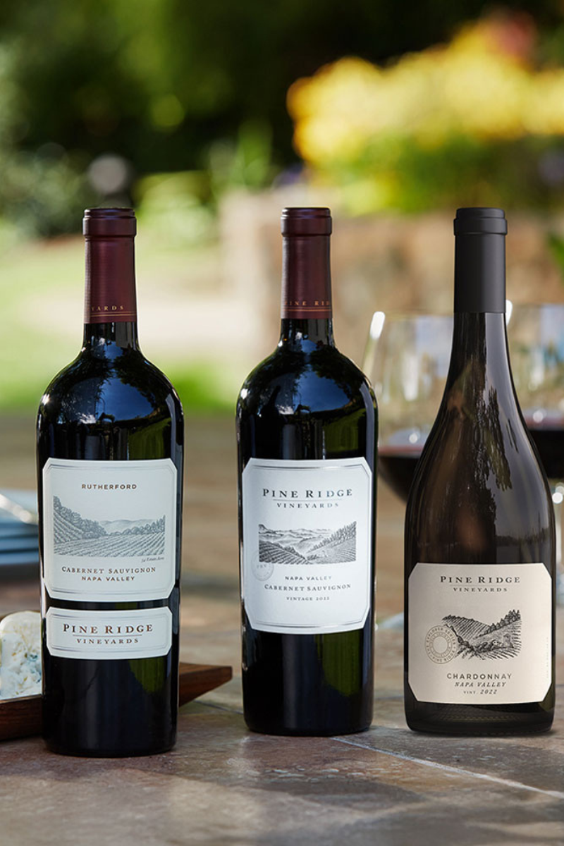Three bottles from the Summer Grilling 3-Bottle Collection—two red Cabernets and one white Chardonnay—are displayed on a table outdoors with wine glasses and blurred greenery in the background.