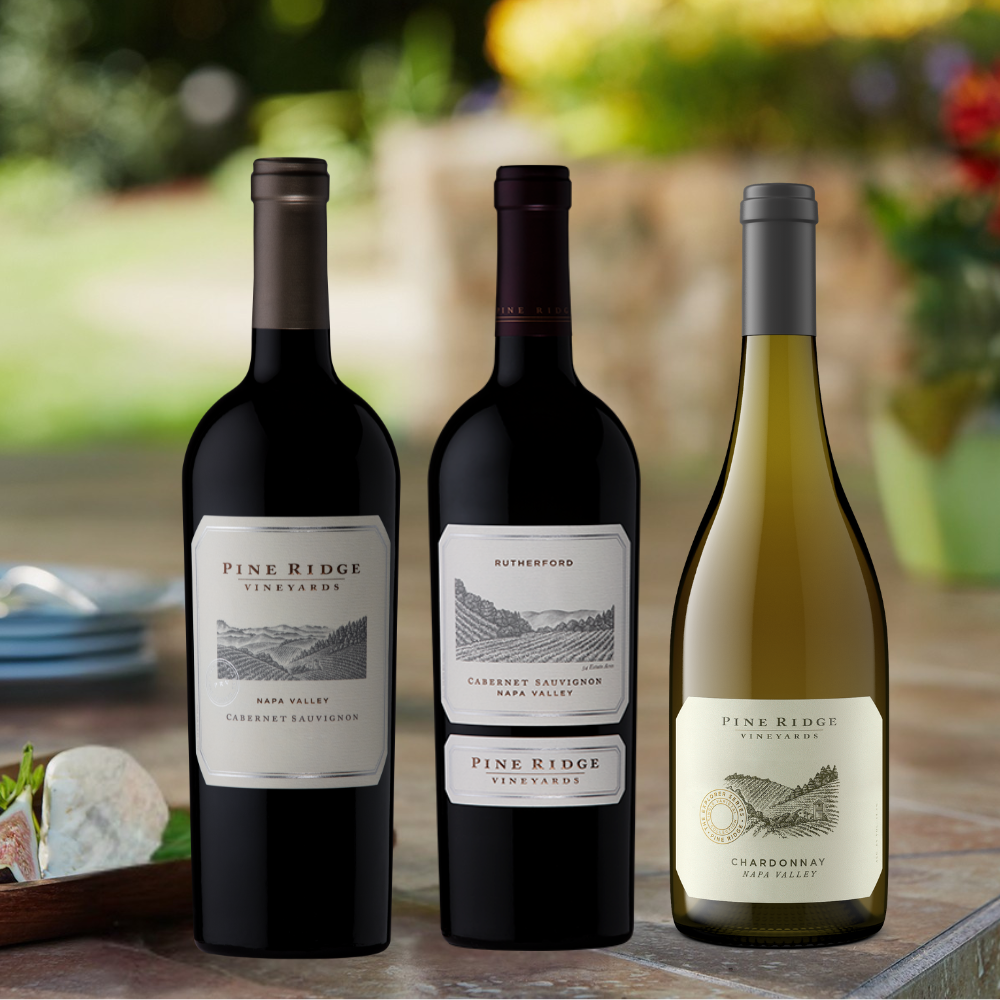 Three bottles of wine from the Summer Grilling 3-Bottle Collection are arranged side by side on a table outdoors. The first and third are Pine Ridge Vineyards, while the middle bottle is labeled Rutherford, set against a blurred garden background.