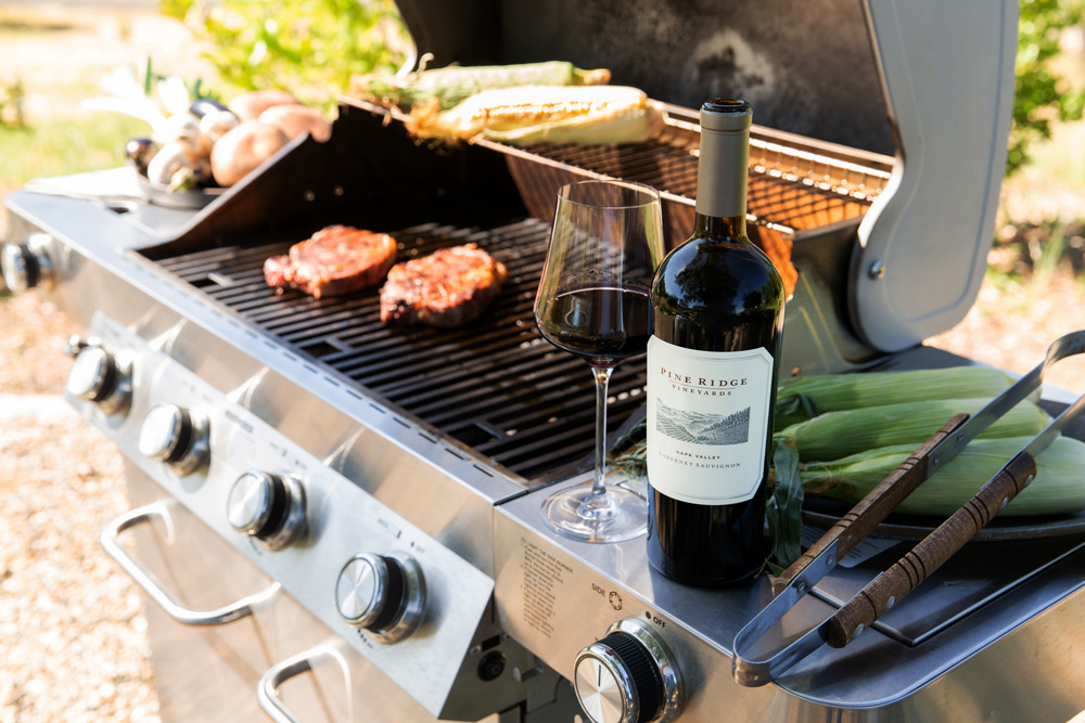 A bottle of red wine and a glass sit on a stainless steel gas grill next to grilling tools; steaks, corn, mushrooms, and other vegetables are cooking on the grill.