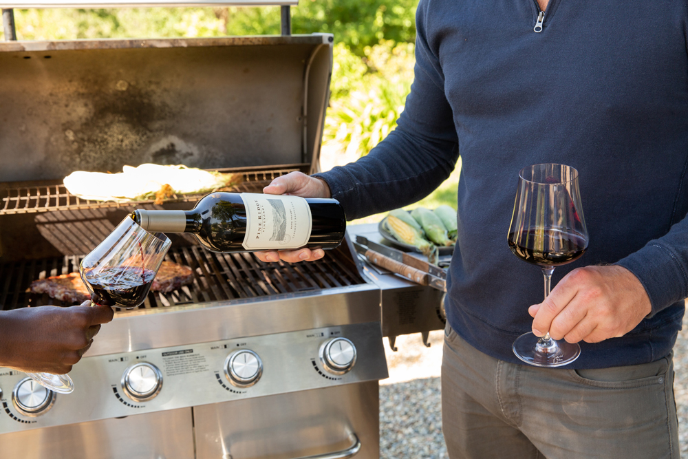 Two people stand in front of an outdoor grill with food cooking, each holding a glass of red wine. One person pours wine from a bottle into the others glass. Grilled vegetables and meat are visible on the grill.