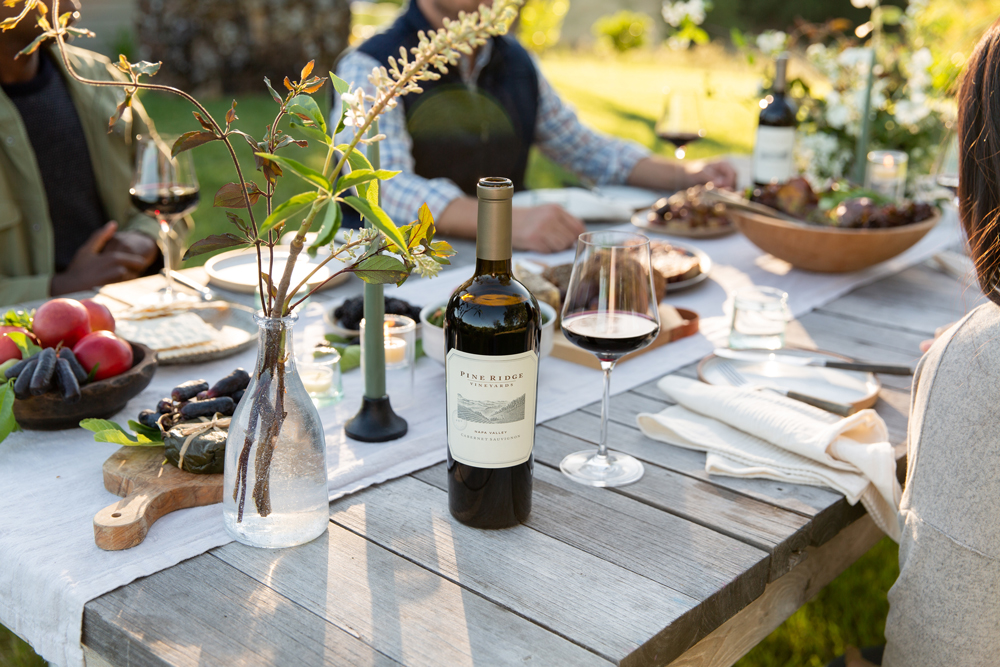 A bottle of Pine Ridge Napa Valley Cabernet and a glass sit on a rustic wooden outdoor dining table set with food, plates, and a vase with branches, with people seated around the table in natural light.