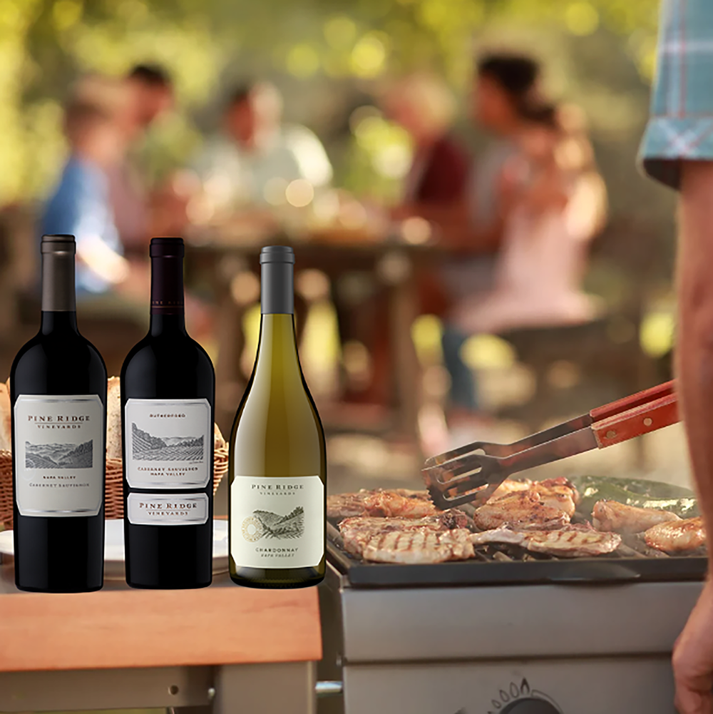 Three bottles of Pine Ridge wine from the 2022 Stags Leap District 3-Bottle Collection are on a table near a barbecue grill sizzling with meat, while friends enjoy time together around a picnic table in a lush garden setting.