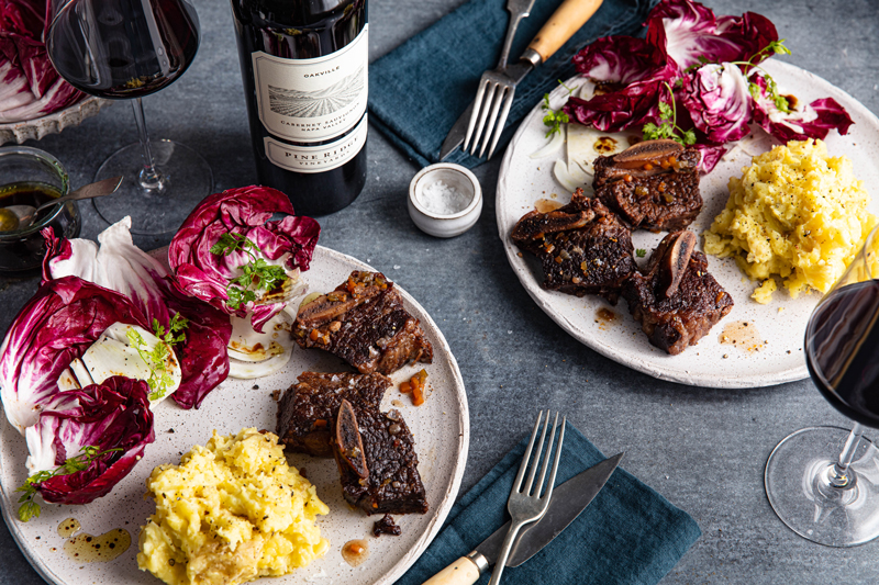 Two plates of food with grilled short ribs, mashed potatoes, and radicchio salad sit on a table. A bottle and glasses of red wine, utensils, and a small bowl of salt are arranged alongside the plates.