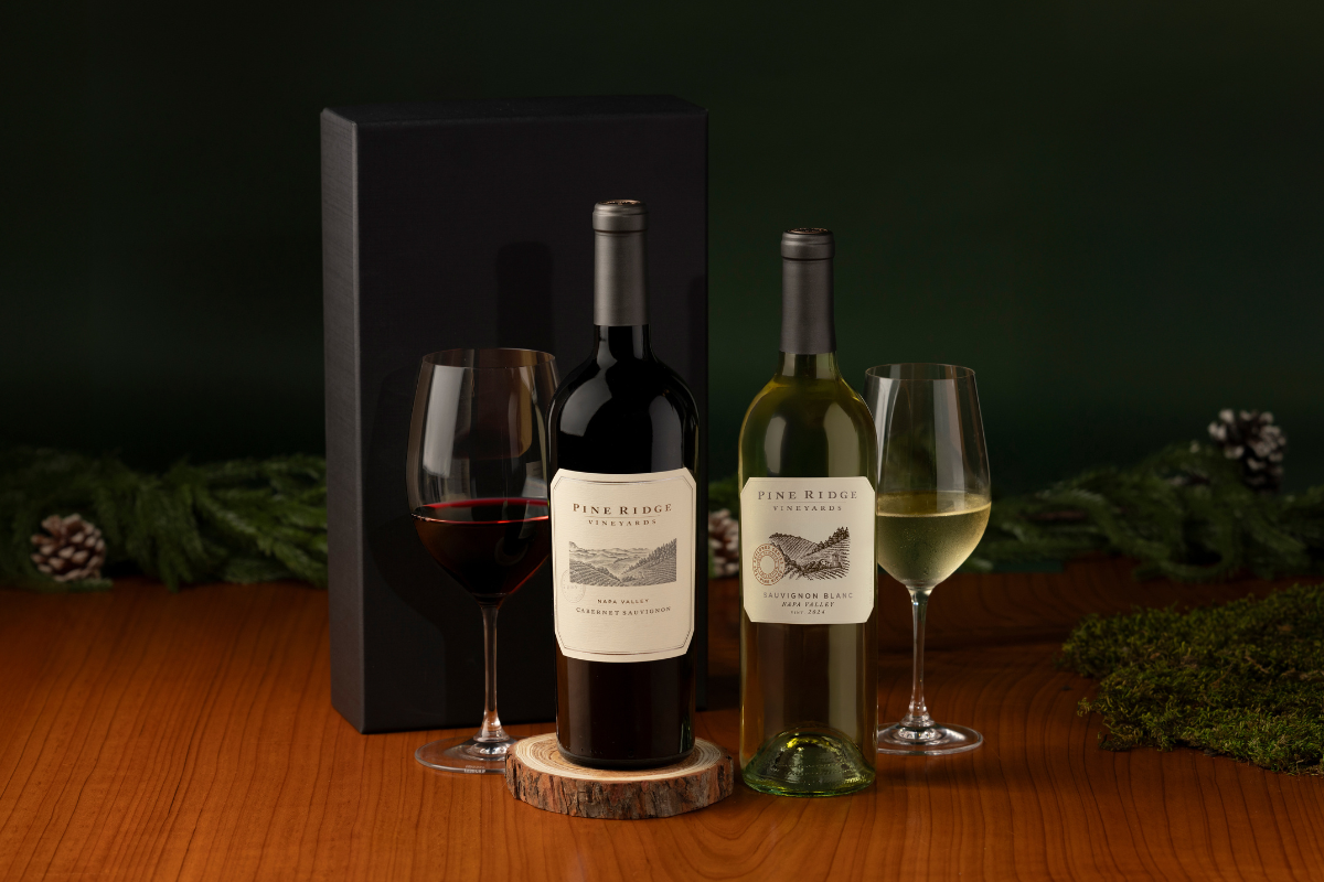 A bottle of red wine and a bottle of white wine stand beside two filled wine glasses on a wooden table. A black gift box and evergreen branches are in the background.