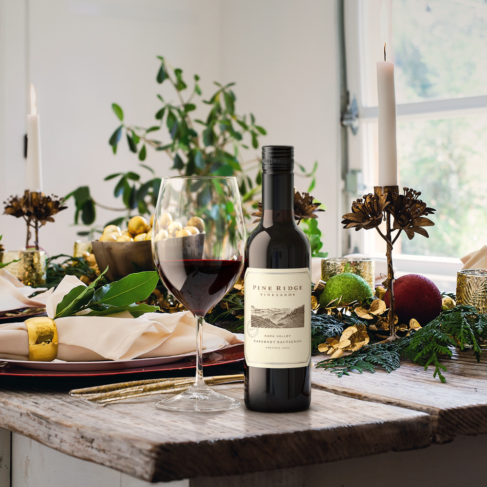 A bottle of Pine Ridge Vineyards 2022 Napa Valley Cabernet Sauvignon and a glass of red wine sit on a rustic wooden table set for a holiday meal, decorated with greenery, candles, and festive ornaments—the perfect addition to any 6-Bottle Collection.