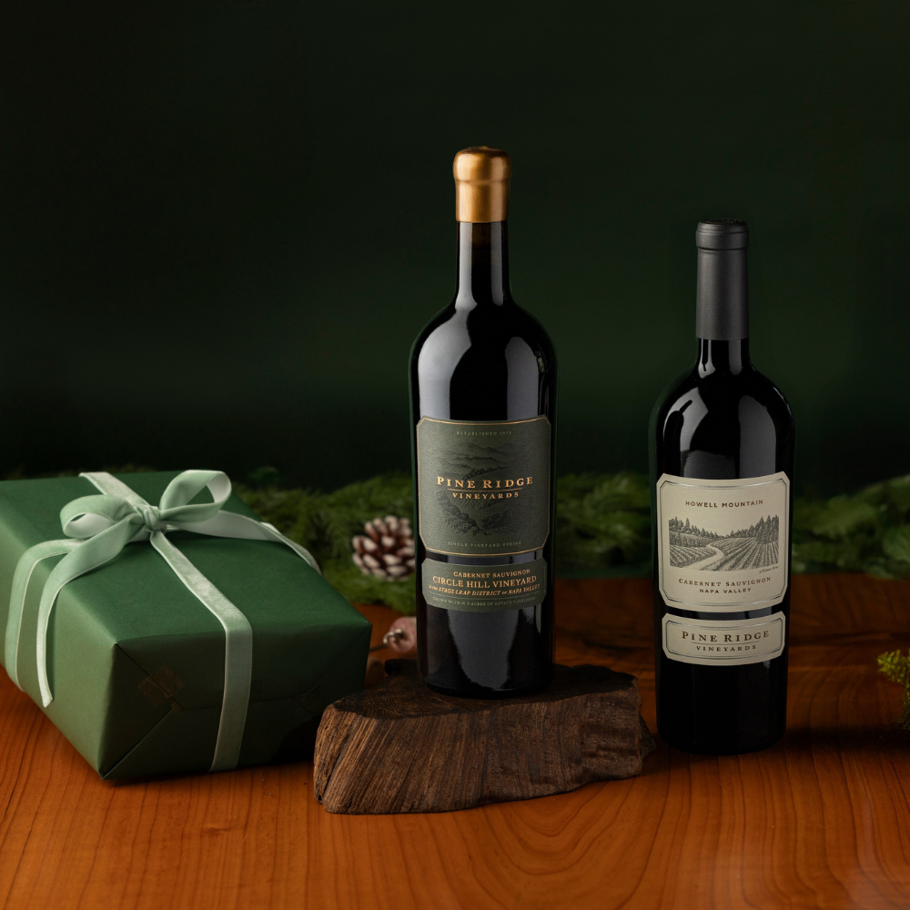 Two bottles of Pine Ridge Vineyards wine, featured in the Napa Valley Favorites 2-Bottle Gift, stand on a wooden surface—one elevated on a block. A green gift box with a white ribbon sits to the left, framed by pine branches in the background.