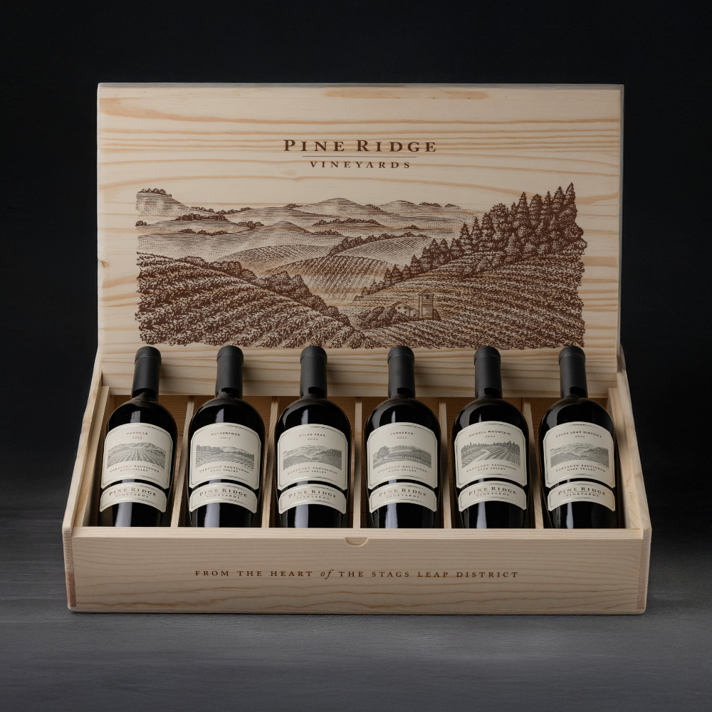 A wooden box with a vineyard illustration on the lid holds the Napa Valley Icons 6-Bottle Collection from Pine Ridge Vineyards, each red wine bottle featuring a white label and black cap, arranged in a single row against a dark background.