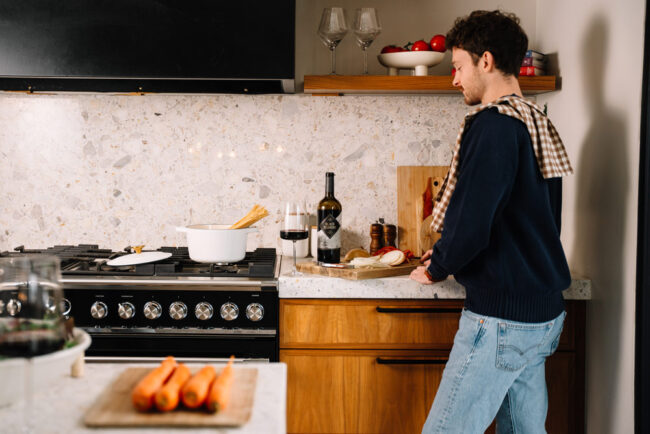 A person stands at a kitchen counter slicing an apple. There is a bottle of wine, two glasses, and a pot of pasta on the stove. Carrots are on a nearby cutting board in the foreground.