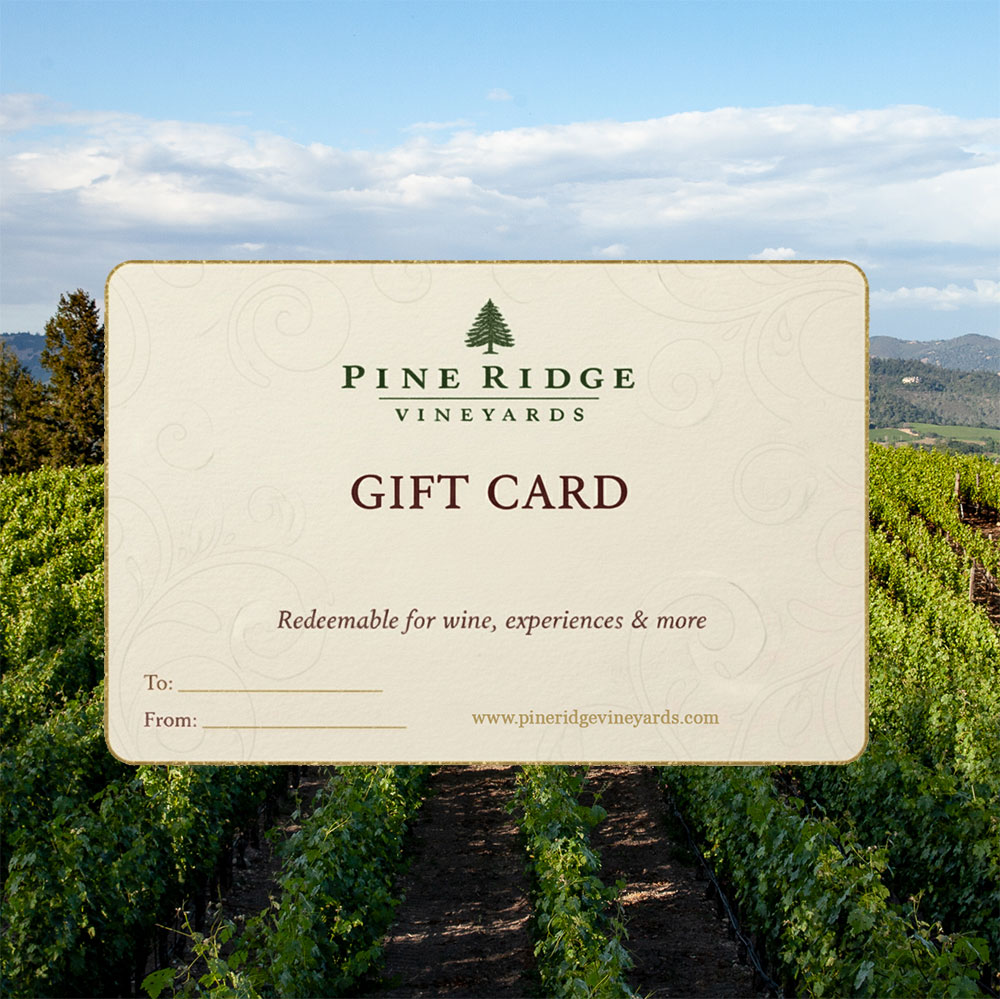 A Pine Ridge Vineyards gift card with fields for To and From is shown in the foreground, with green vineyard rows and hills under a partly cloudy sky in the background.