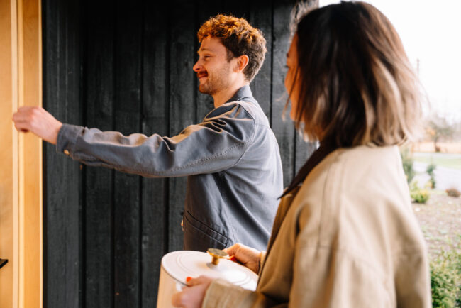 A man in a gray jacket knocks on a door while a woman in a beige coat stands beside him, holding a white pot with a lid. They are outside near a dark wooden building.