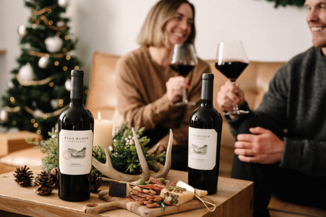 Two bottles of red wine are on a table with a cheese and nut platter, pinecones, and a lit candle. In the background, a smiling man and woman sit on a couch, clinking wine glasses. A decorated Christmas tree is visible.