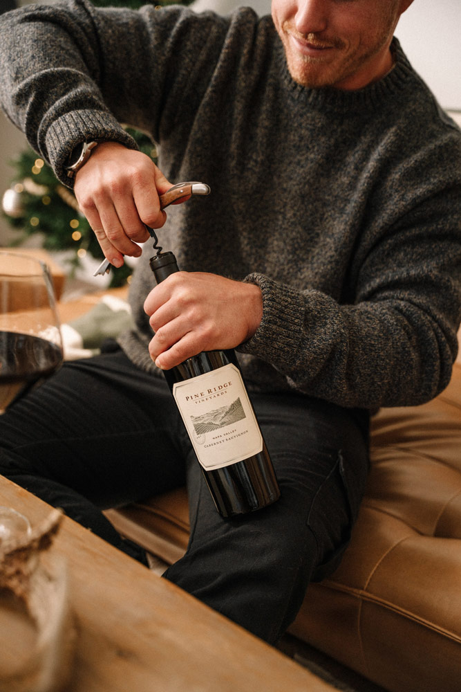 A person wearing a dark sweater sits on a tan couch, using a corkscrew to open a bottle of red wine. A large glass of red wine is on a wooden table in the foreground.
