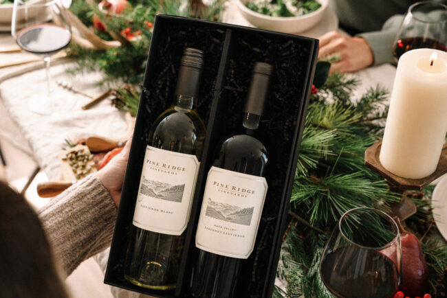 A person holds an open box containing two bottles of Pine Ridge Vineyards wine on a festive table set with greenery, candles, glasses of wine, and food.