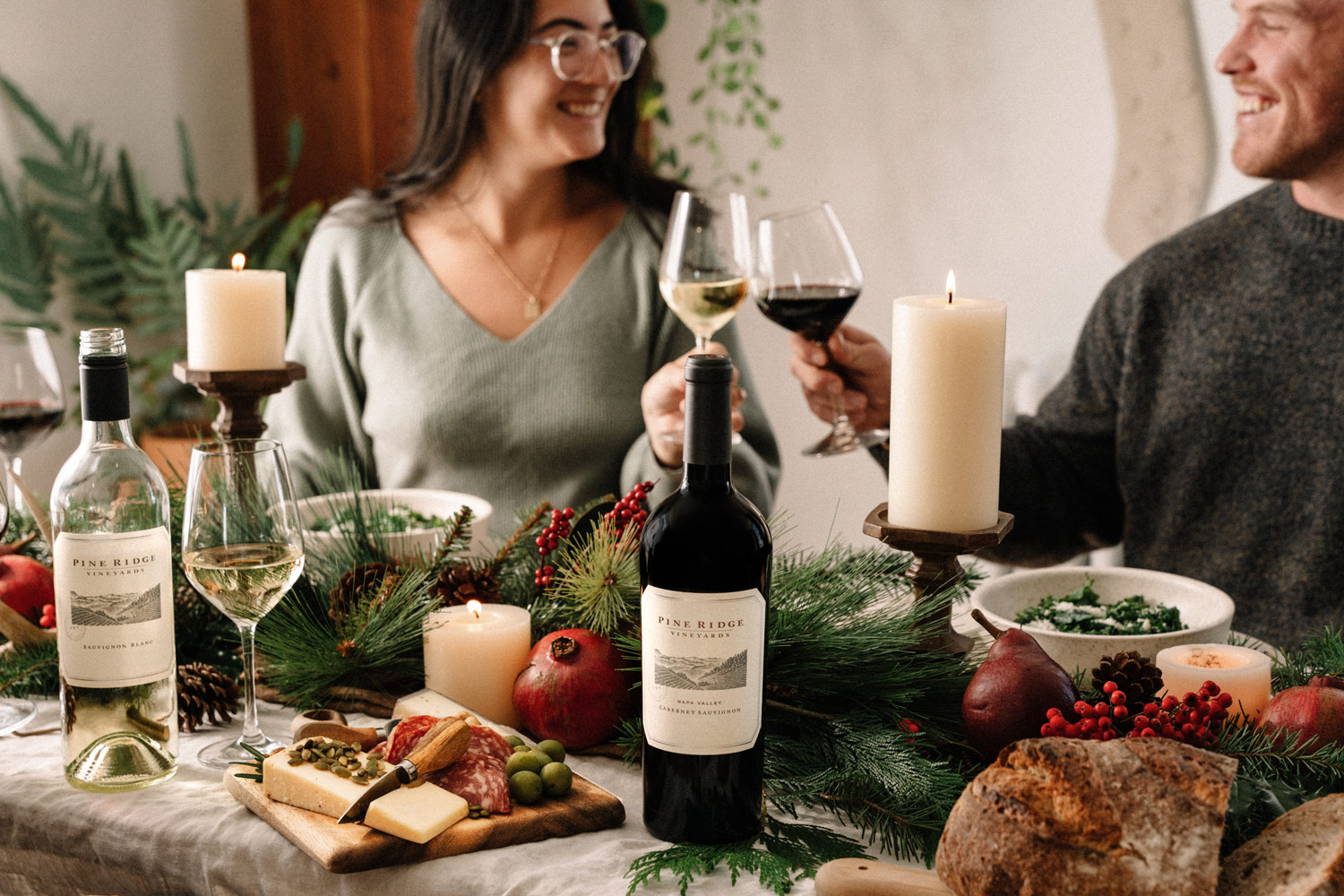 Two people smile and clink wine glasses at a decorated dining table with candles, wine bottles, cheese, bread, fruit, and greenery. The atmosphere appears festive and cozy.