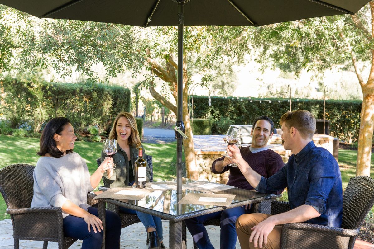Four people sit around a square outdoor table under a black umbrella, holding wine glasses and smiling. There are two wine bottles and glasses on the table, with trees and greenery in the background.