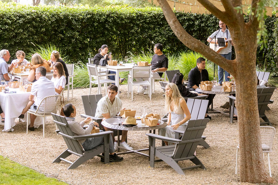 People are dining at outdoor tables on a gravel patio surrounded by greenery. Some enjoy Adirondack chairs while a musician plays guitar nearby. The relaxed atmosphere is perfect for savoring Wine Club Perks with friends.