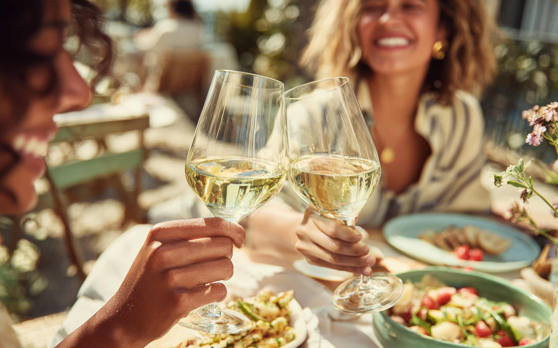 Two people clink glasses of refreshing white wines while sitting at an outdoor table set with plates of salad and pasta on a sunny day, both smiling and enjoying the meal.