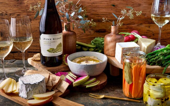 A rustic tabletop displays wine glasses, a bottle of refreshing white wine, assorted cheeses, crackers, sliced apples, dip with vegetables, pickled vegetables in jars, and wooden vases with greenery against a wooden backdrop.