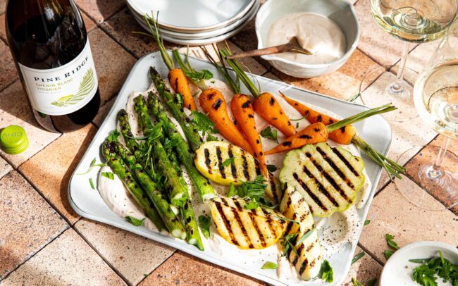 A tray of grilled vegetables, including asparagus, carrots, and sliced halloumi cheese, garnished with herbs and served with a white dipping sauce. Nearby are wine glasses, plates, and a bottle of refreshing white wine.