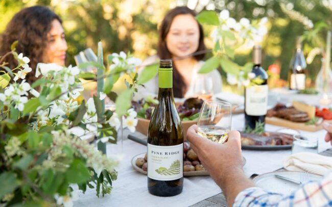 Three people sit at an outdoor table with food, flowers, and two bottles of wine. One person holds a glass, focusing on a bottle labeled “Pine Ridge”—perfect for enjoying refreshing white wines in the bright, green garden setting.