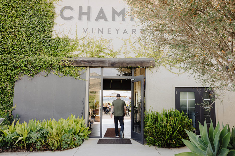 A person walks into the entrance of a building partially covered in greenery and vines, with the word VINEYARD above the doorway—an inviting spot for those seeking Wine Clubs with Great Perks amid lush plants and trees.
