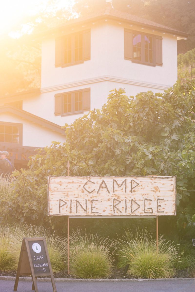 A wooden sign reading Camp Pine Ridge stands in front of green bushes and tall grass, with a white two-story building and a small registration sign visible in the background under bright sunlight.