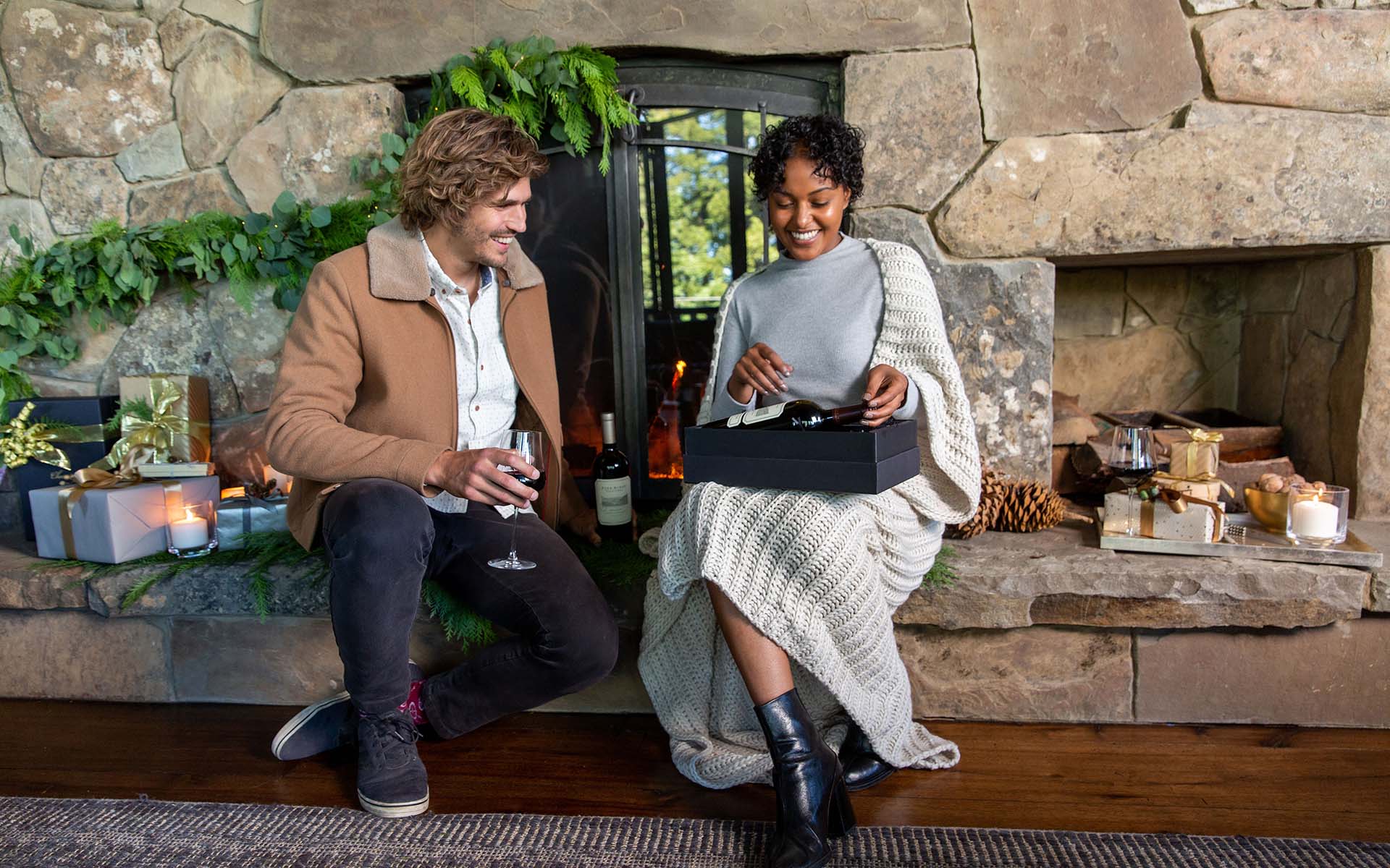 A man and woman sit by a stone fireplace surrounded by candles and wrapped gifts. The woman opens a black holiday wine gift box while the man holds a glass of wine. Both smile warmly, dressed in cozy, casual clothing.