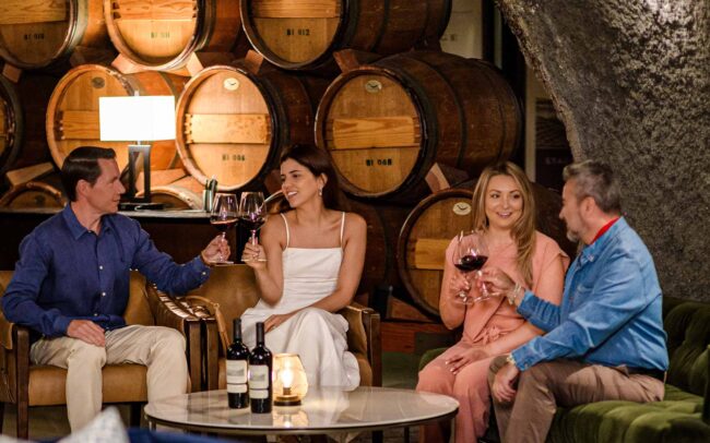 Four people sit on sofas in a wine cellar, holding and clinking glasses of red wine. Wooden barrels are stacked in the background, and a candle and wine bottles are on the glass coffee table in front of them.