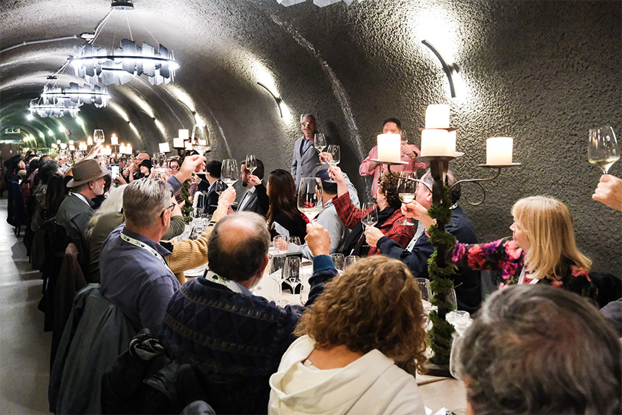 A large group of people seated at long tables in a tunnel-like room raise their glasses in a toast, reminiscent of Wine Clubs with Great Perks. The festive atmosphere is enhanced by plates, glasses, and candles adorning the tables.