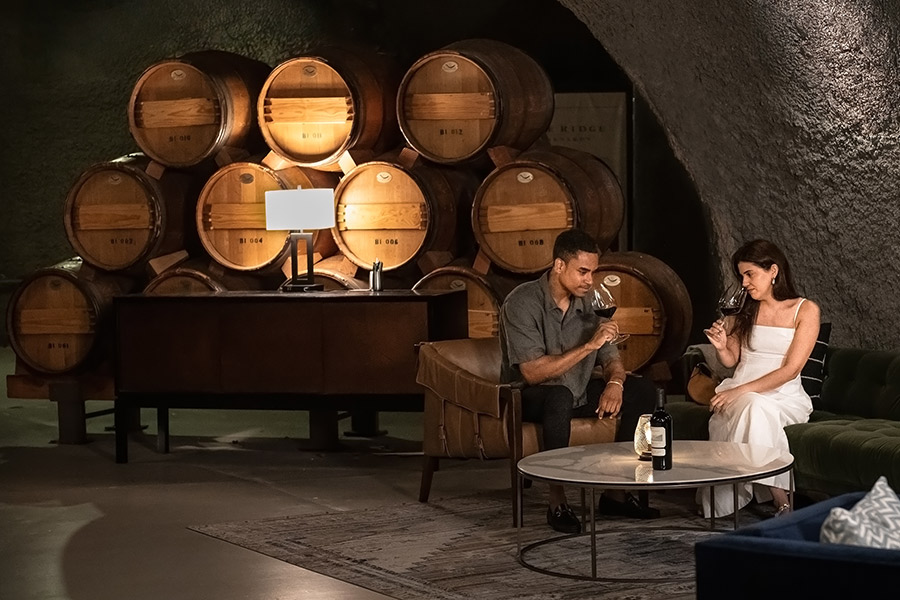 A man and a woman sit on sofas in a wine cellar, holding glasses of wine. Behind them are stacked wooden barrels. The relaxed, dimly lit scene feels perfect for discussing Wine Clubs with Great Perks over a bottle by the lamp-lit table.