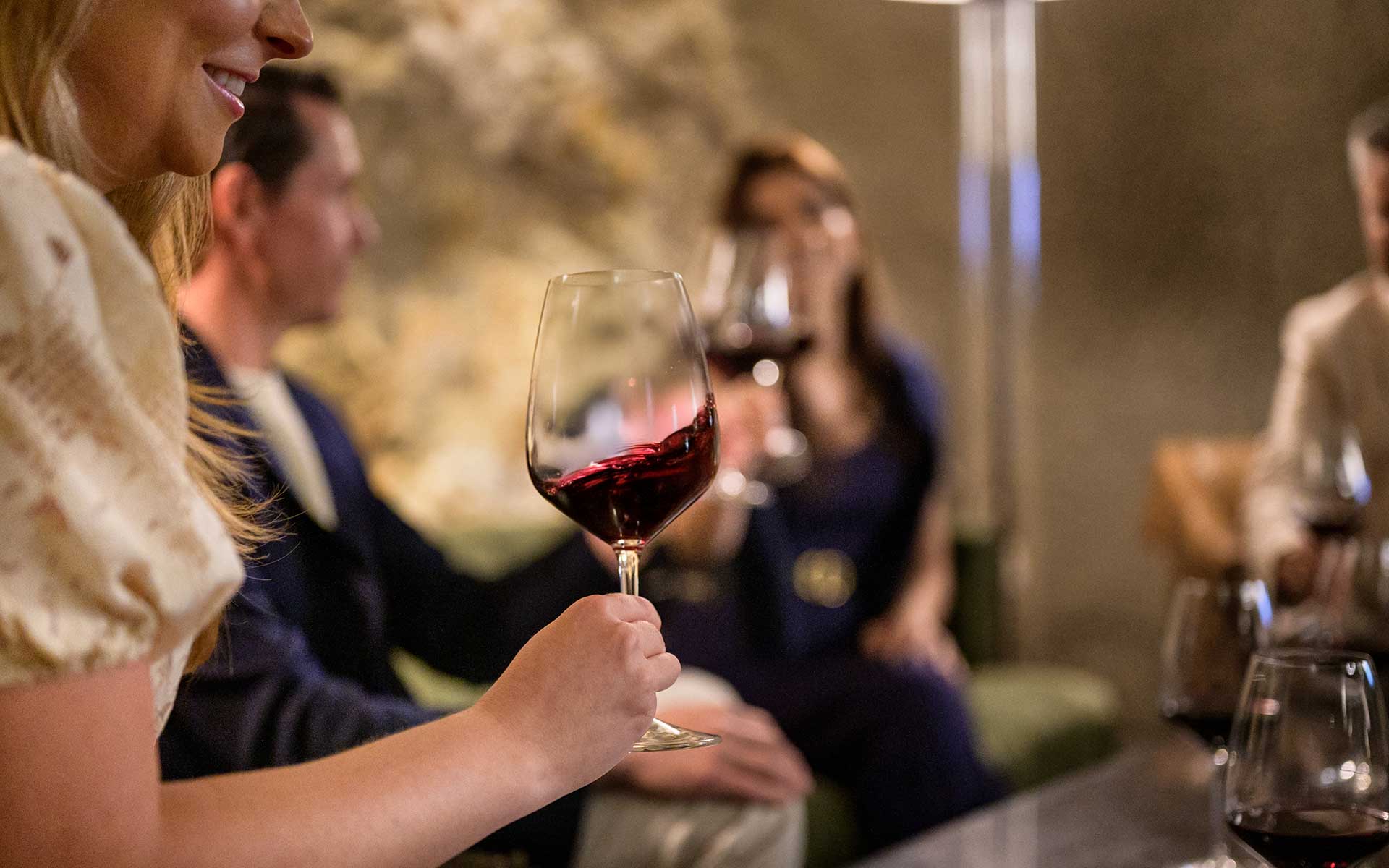 A group of people sits on a couch in a dimly lit room, each holding a glass of red wine. The focus is on a woman in the foreground holding her glass, while others are blurred in the background.