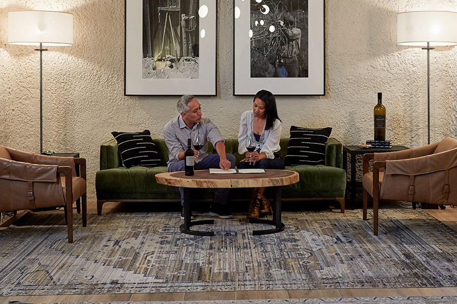 A man and woman sit on a green couch in a modern living room, sharing a bottle from one of the Wine Clubs with Great Perks. Two leather chairs, a large rug, lamps, and framed artwork create an inviting atmosphere behind them.