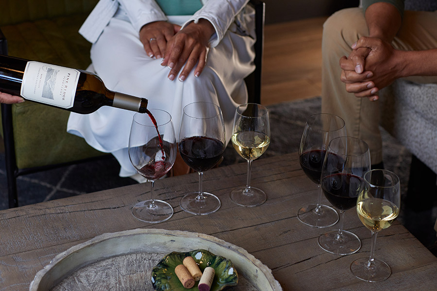 A person pours red wine into a glass on a table with several glasses of red and white wine—reminiscent of gatherings hosted by Wine Clubs with Great Perks. Two people sit nearby, hands visible, and a tray holds corks and small items.