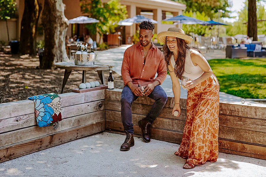 A man sits on a wooden bench while a woman in a sun hat stands nearby, holding a ball and smiling, enjoying an afternoon with friends, drinks from Wine Clubs with Great Perks, and bocce balls scattered under the trees.