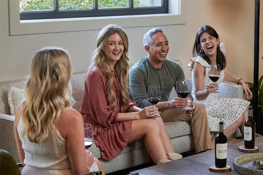 Four people sit on a couch and chairs indoors, smiling and holding glasses of red wine. Enjoying Wine Club Perks, they have two bottles of wine and a small dish on the coffee table. A window is visible in the background.