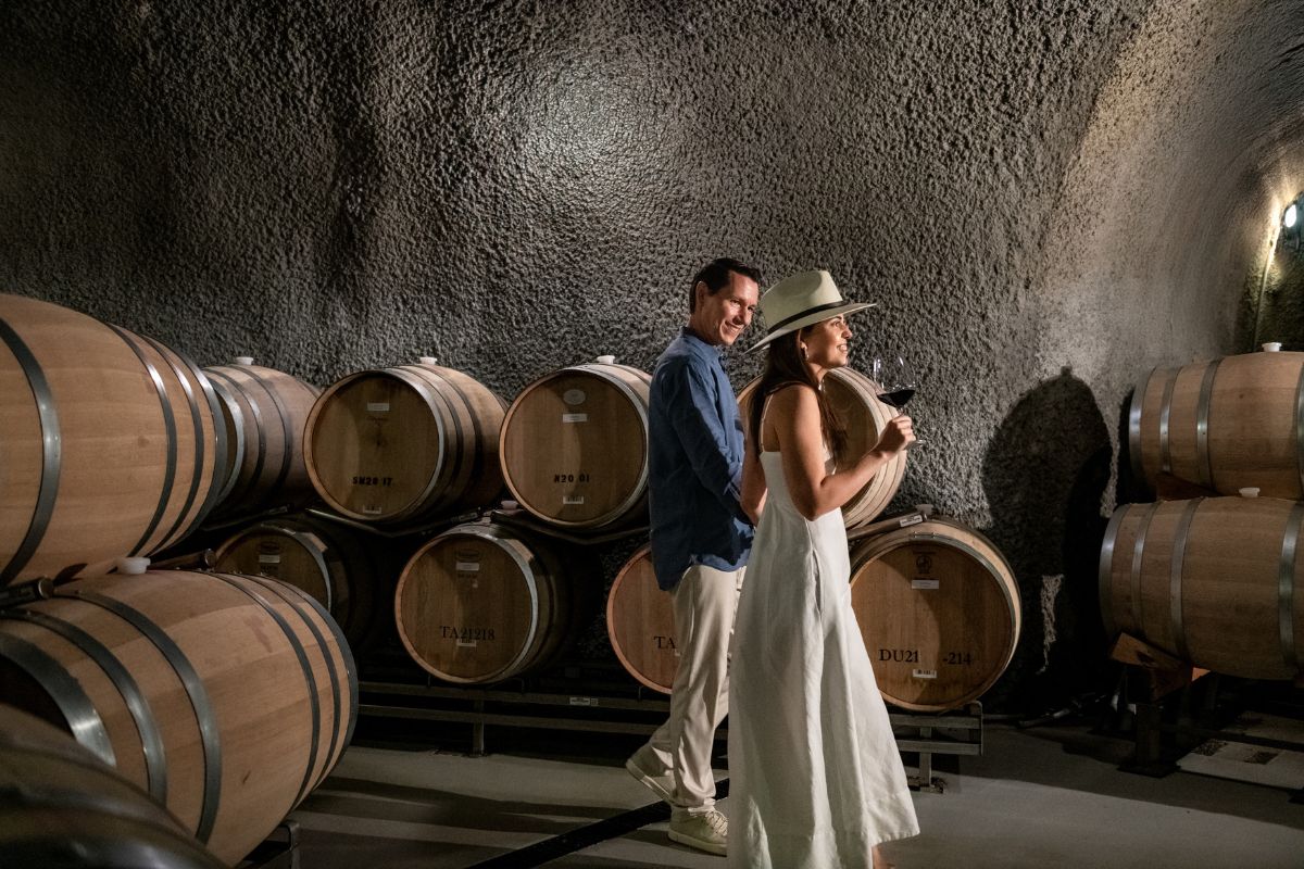 A man and a woman stroll through a wine cellar lined with wooden barrels, celebrating Noon Year’s Eve. She holds a glass of red wine, dressed in a white dress and hat; he walks beside her in a blue shirt and white pants.