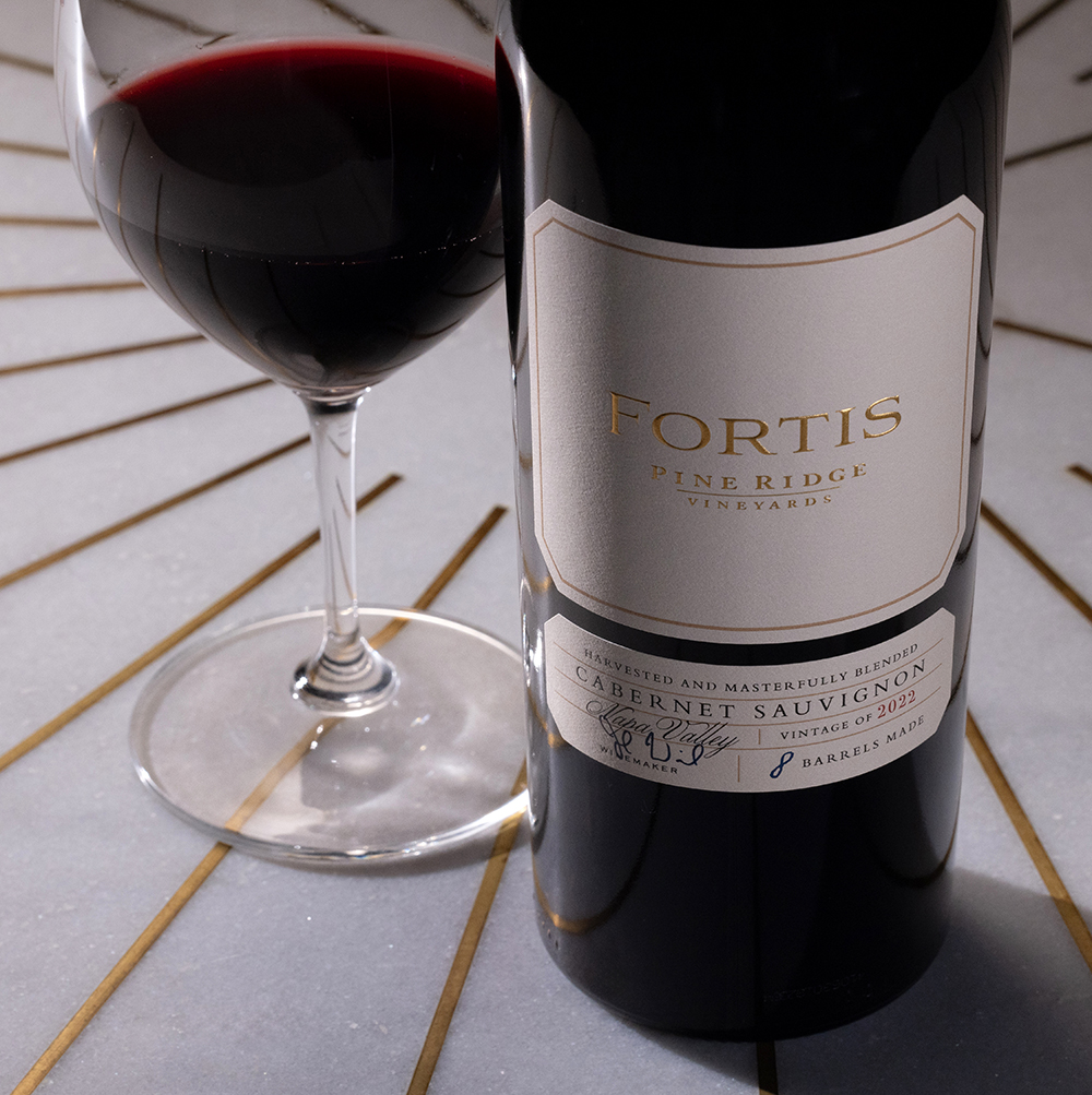 A bottle of Fortis Pineridge Vineyard Cabernet Sauvignon 2021 is placed next to a glass filled with red wine on a white surface with gold lines. The bottle label is clearly visible.